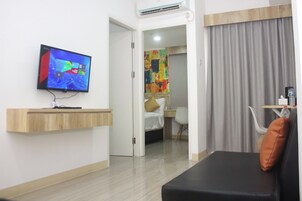 Quarto de hotel com televisão de tela plana na parede, uma estante de madeira, cama com cabeceira branca, escrivaninha com cadeira e uma grande janela com cortinas.