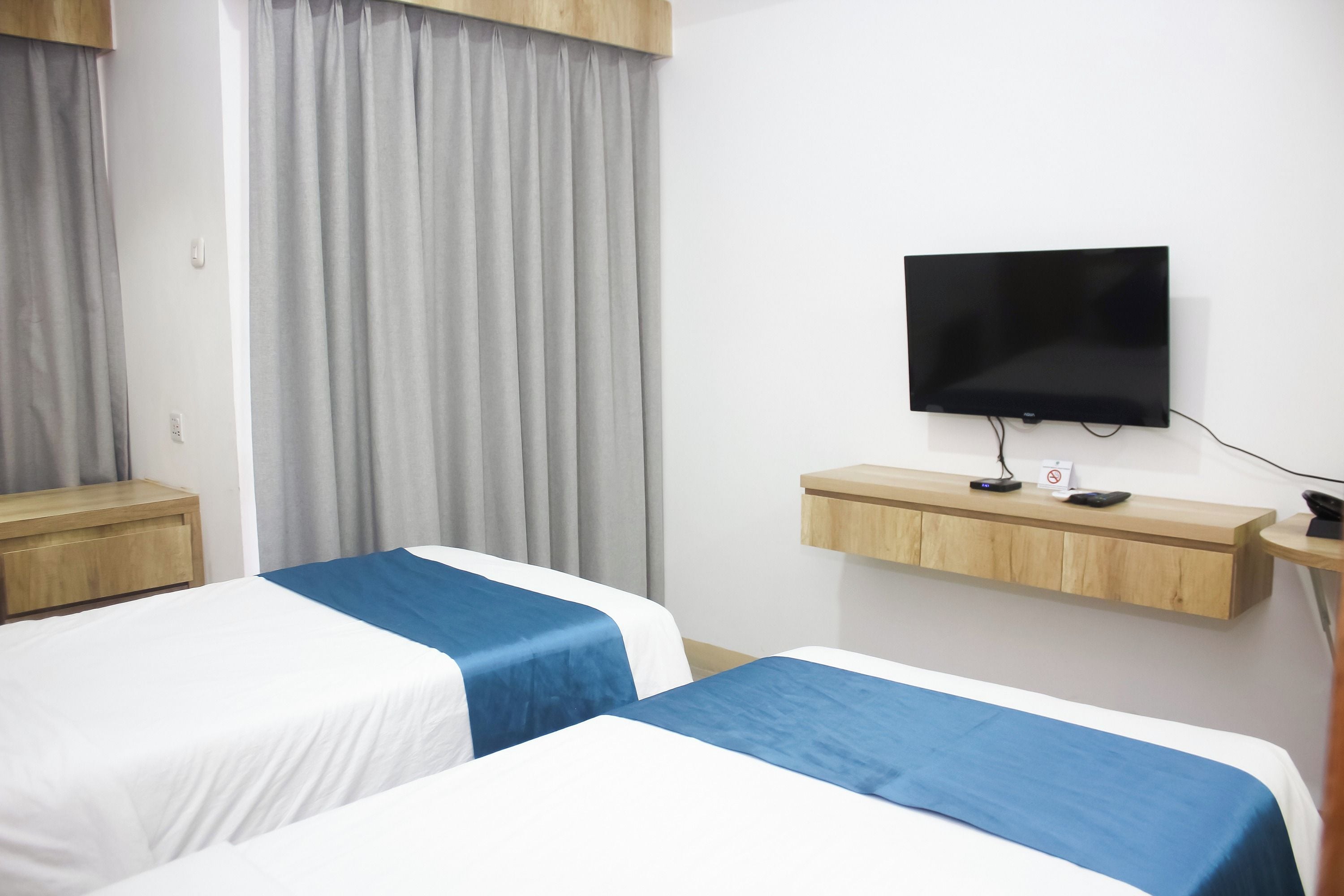 Habitación de hotel con dos camas, un televisor en un estante de madera, y un escritorio con un teléfono y un cartel.