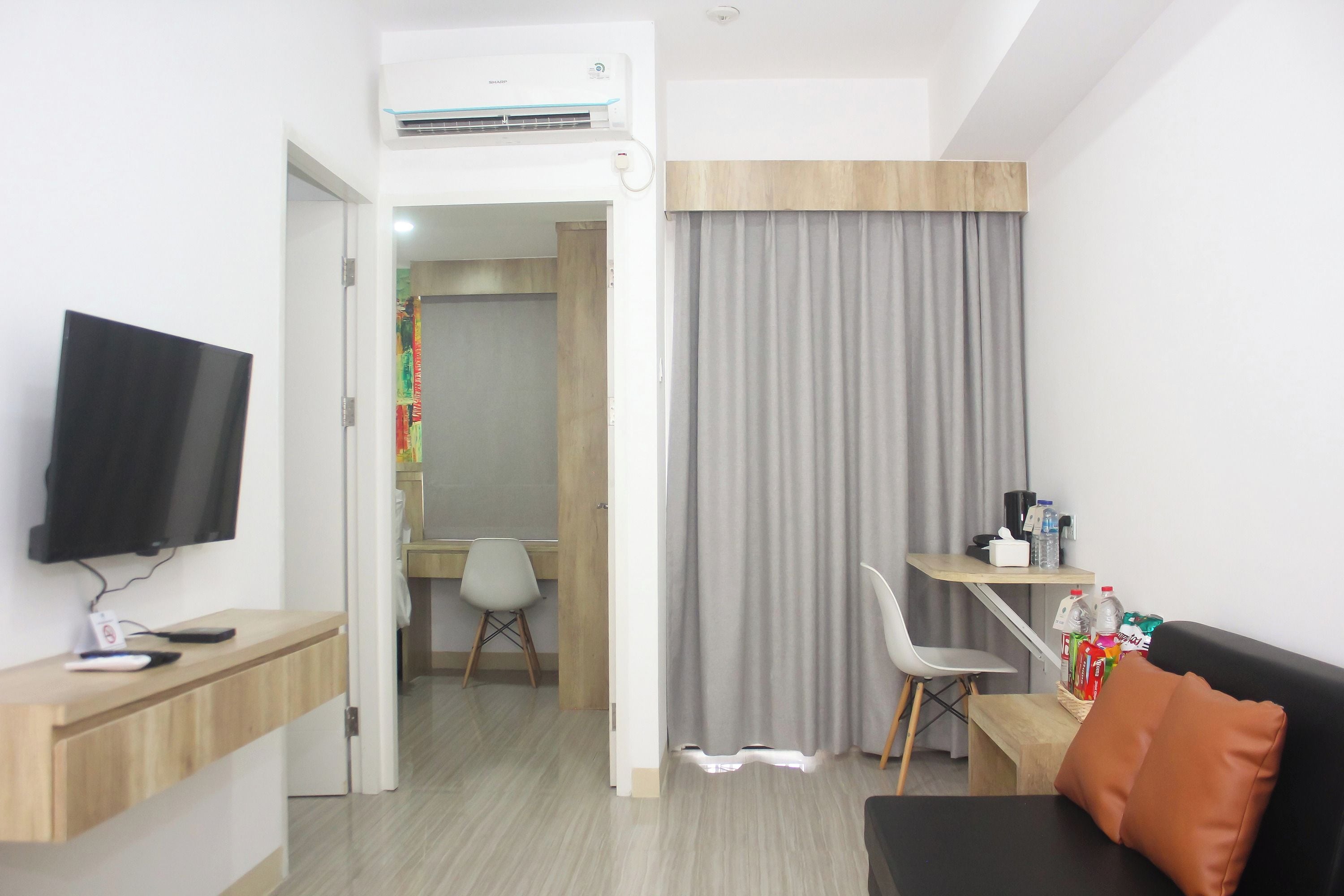 Een moderne hotelkamer met een flatscreen televisie, een houten bureau, een zwarte bank met oranje kussen, een grijze gordijn en een klein tafeltje met water en snacks.