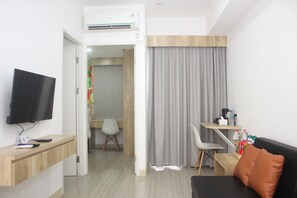 Een moderne hotelkamer met een flatscreen televisie, een houten bureau, een zwarte bank met oranje kussen, een grijze gordijn en een klein tafeltje met water en snacks.