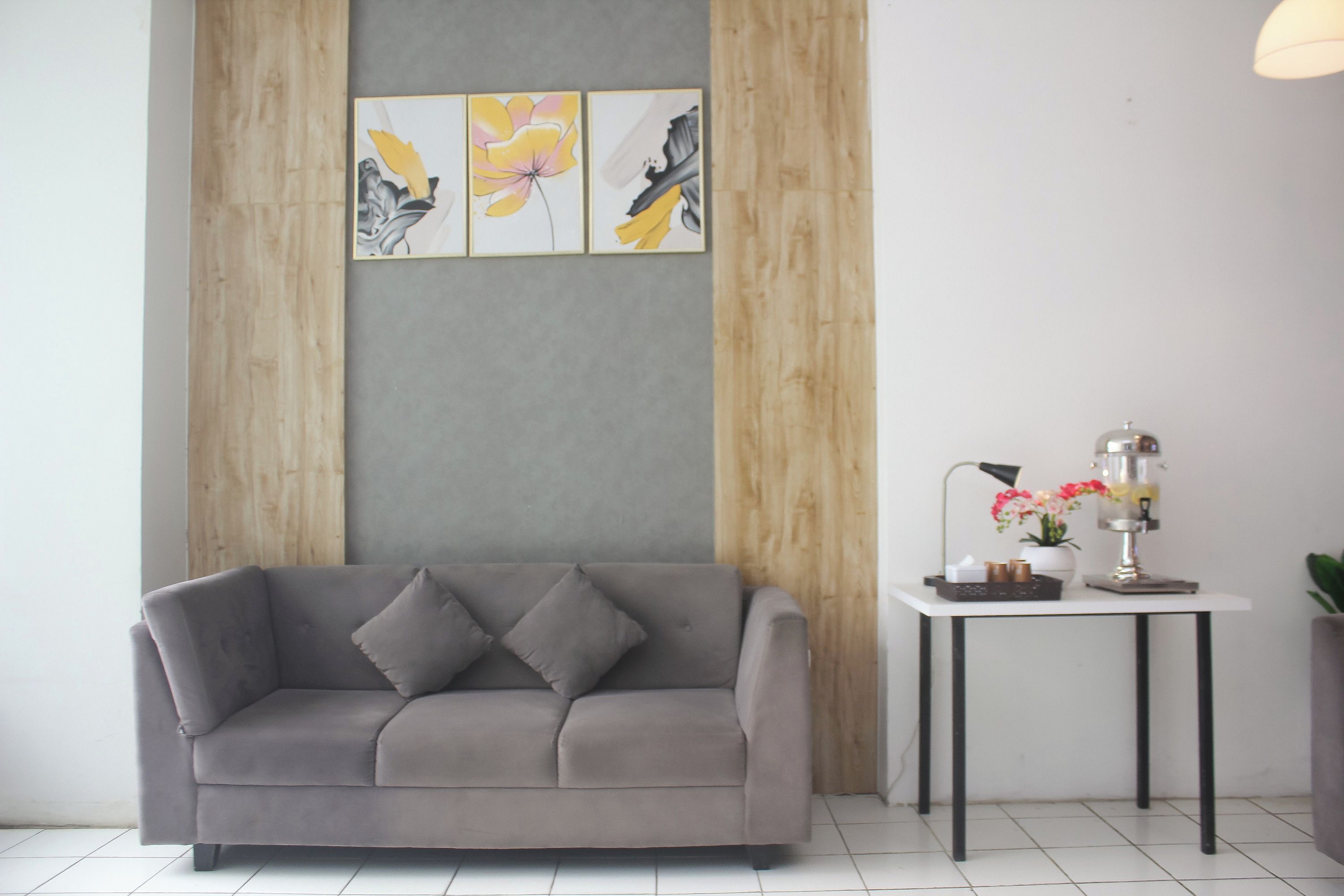 Ein modernes Wohnzimmer mit einem grauen Sofa, einem kleinen Tisch mit Lampe und Blumen sowie drei gerahmten Kunstwerken an der Wand.
