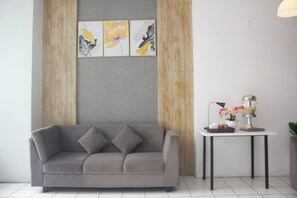 Ein modernes Wohnzimmer mit einem grauen Sofa, einem kleinen Tisch mit Lampe und Blumen sowie drei gerahmten Kunstwerken an der Wand.