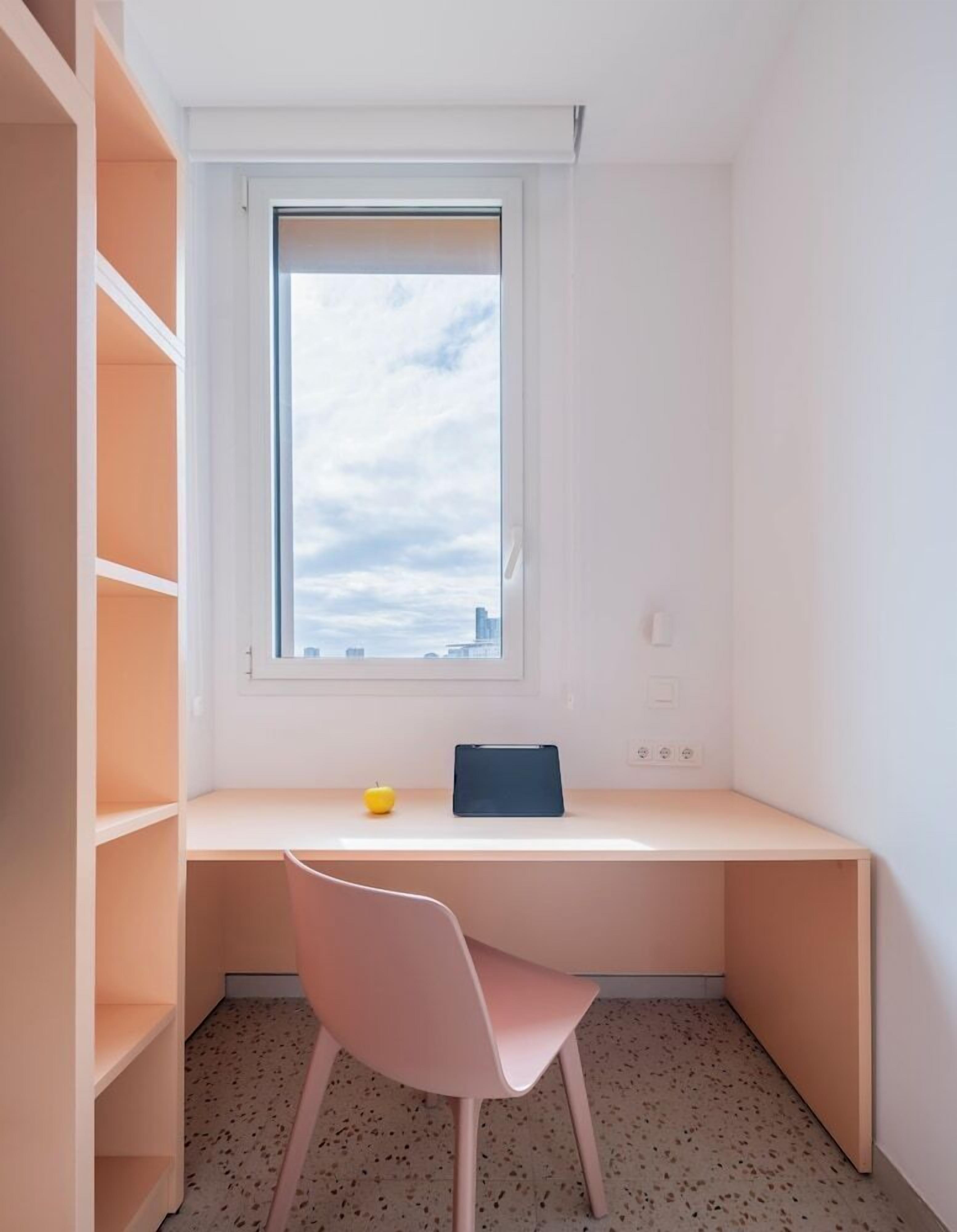 Una habitación pequeña y luminosa con escritorio, silla y ventana con vistas a un paisaje urbano.