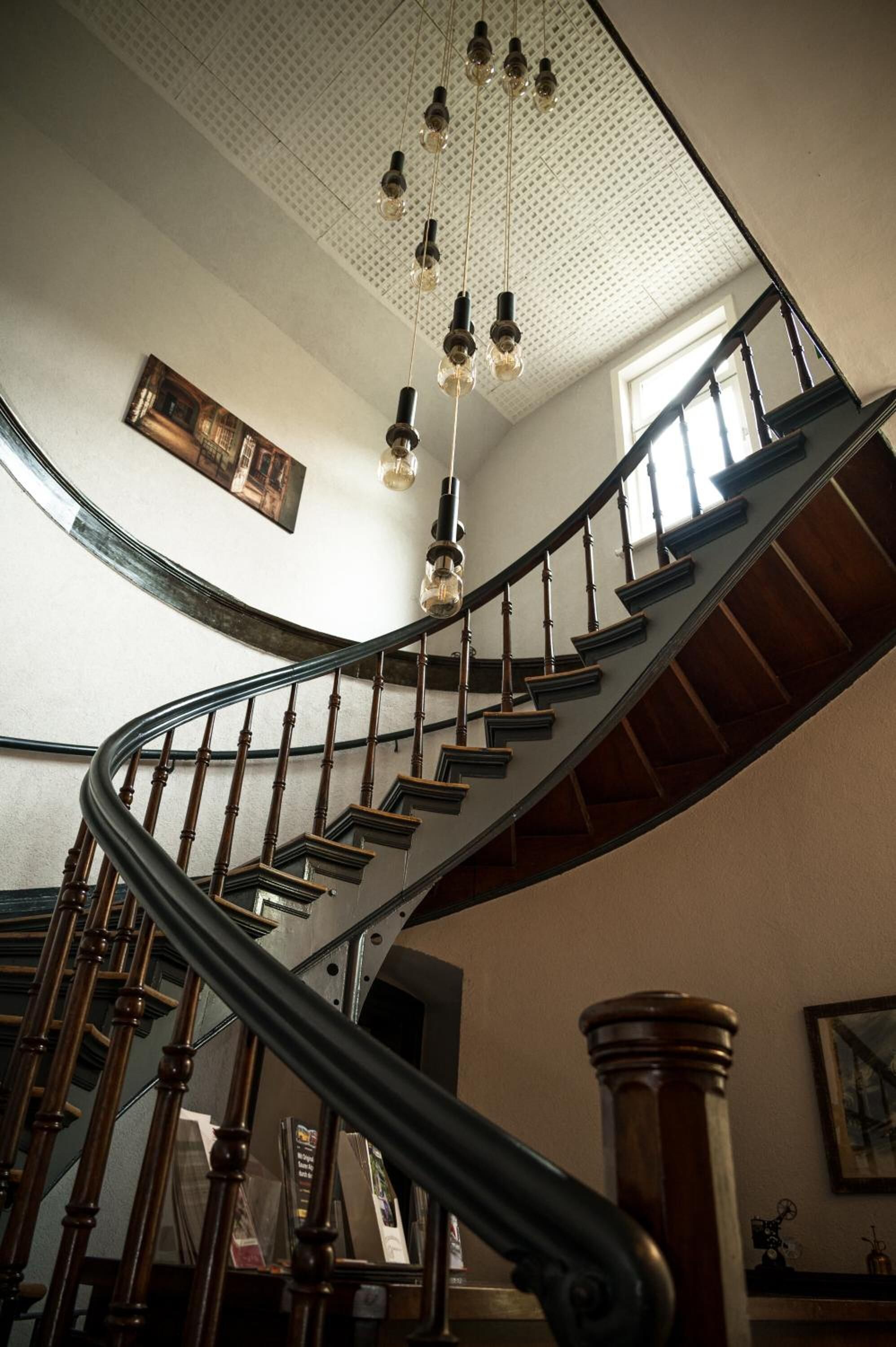 Eine geschwungene Holztreppe mit Metallgeländer, eine Decke mit hängenden Leuchten und ein gerahmtes Bild an der Wand.