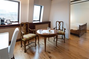 Una habitación con piso de madera, dos sillas de madera con cojines a rayas, una mesita redonda con una taza y un platillo, una cama con ropa blanca y un escritorio con una computadora y una ventana.