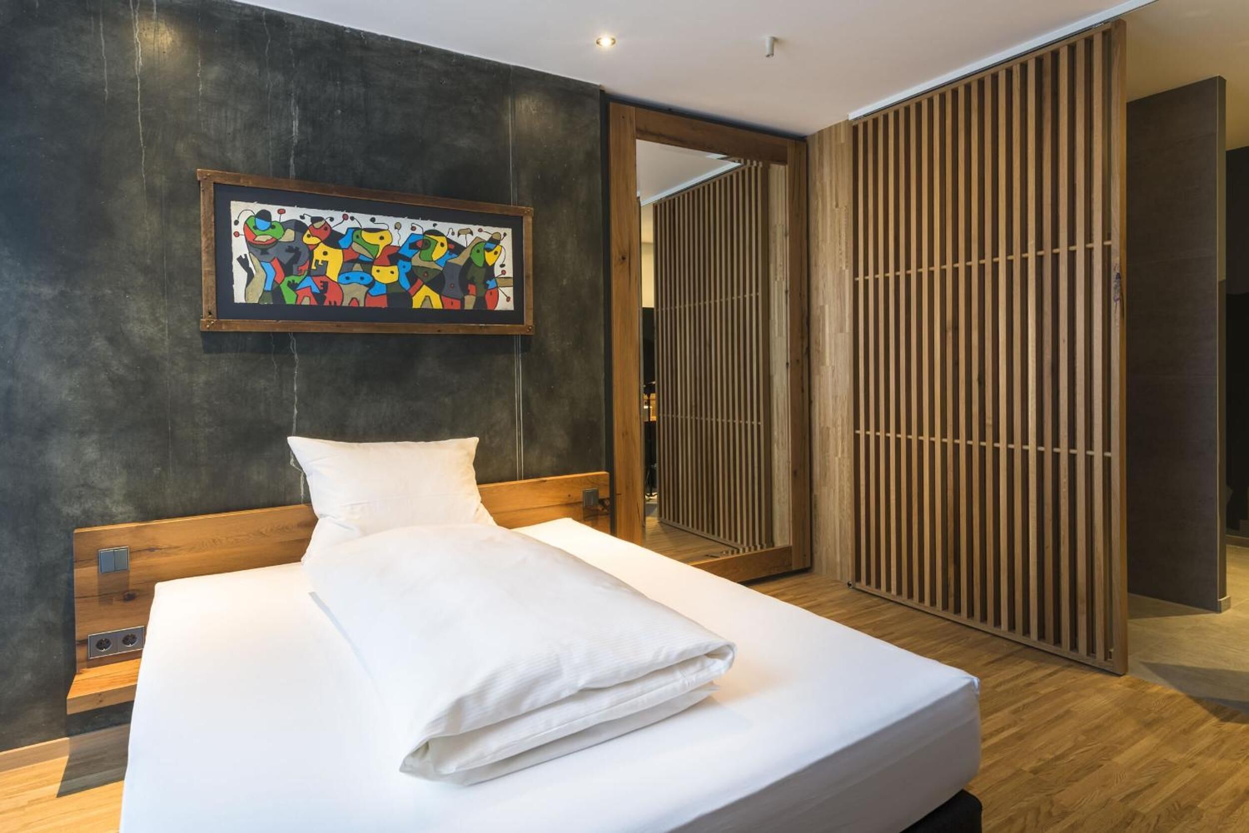 Une chambre d’hôtel moderne avec un grand lit, une tête de lit en bois, un tableau abstrait encadré et une porte coulissante en bois.