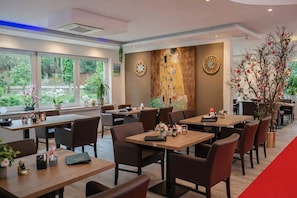 A modern dining area with tables and chairs, a large mural on the wall, and a view of greenery outside. - Hotel Radevormwald (Radevormwald)