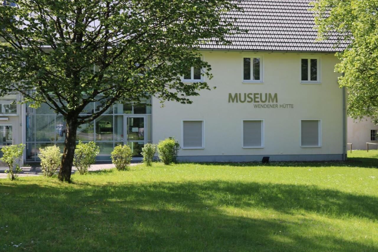 Een gebouw met een betegeld dak, aangeduid met 'MUSEUM WENDENER HÜTTE', omringd door gras en een boom.