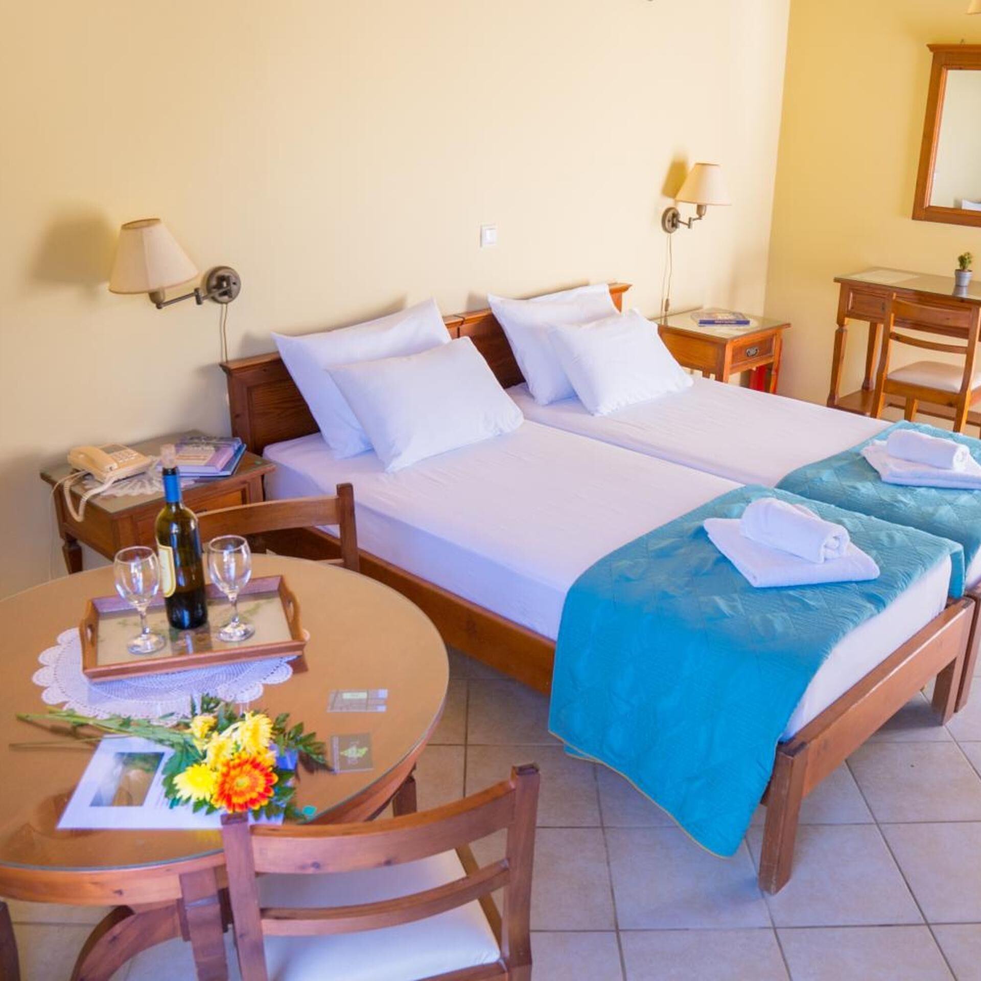 A hotel room with a double bed, a bedside table with a lamp, a small desk with a chair, and a round table with a bottle of wine, glasses, and a flower arrangement.