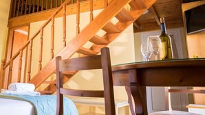 Eine Holztreppe führt zu einer oberen Ebene, ein Esstisch mit einer Flasche und Gläsern sowie ein Bett mit einer blauen Decke.