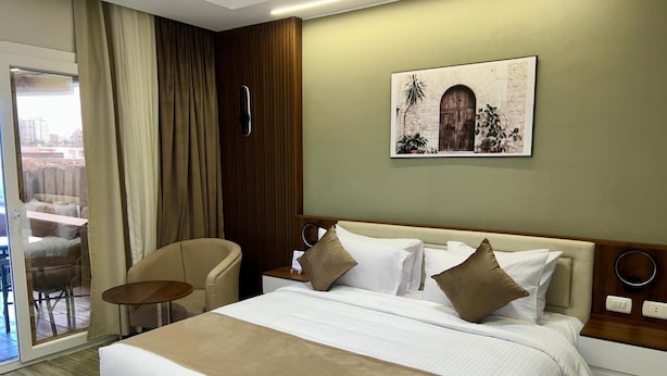A hotel room with a large bed, a chair, a small table, and a view of the city through the window. - XU San Stefano Hotel Alexandria (Alexandria)
