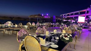An outdoor dining area with tables set for a meal, surrounded by thatched umbrellas and a building with illuminated signage. - XU San Stefano Hotel Alexandria (Alexandria)