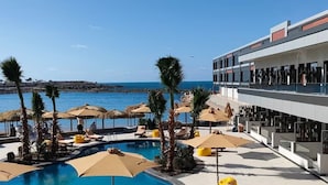 A pool area with lounge chairs and umbrellas, a clear view of the sea, and a modern building with balconies. - XU San Stefano Hotel Alexandria (Alexandria)