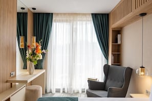A modern hotel room with a bed, a grey armchair, a desk with a vase of flowers, a bookshelf, and a large window with sheer curtains. - Harmony Suites 11, 12 Grand Resort (Nessebar)