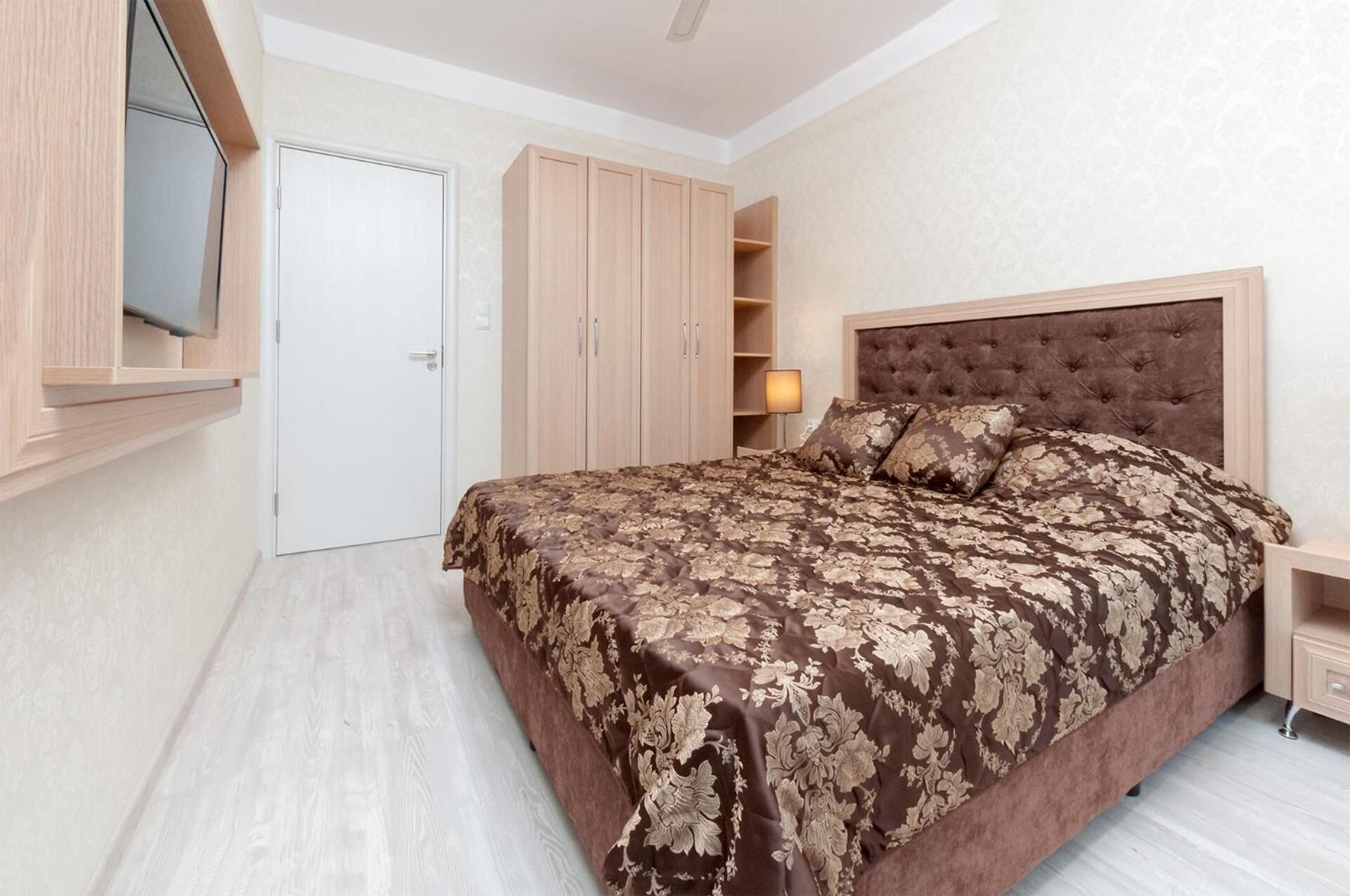 A bedroom with a large bed, a wardrobe, a TV mounted on the wall, and a bedside table with a lamp.