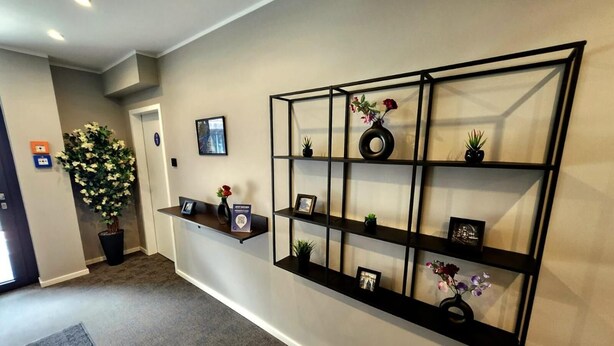 Un área de recepción con un mostrador, una planta grande en maceta, una estantería de pared con objetos decorativos y un cuadro en la pared.