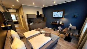 Una habitación de hotel moderna con sofá cama, zona de comedor, cocina y televisor montado en la pared.