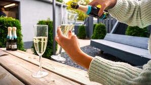 Eine Person schwenkt Champagner in ein Glas auf einem Tisch im Freien aus Holz.