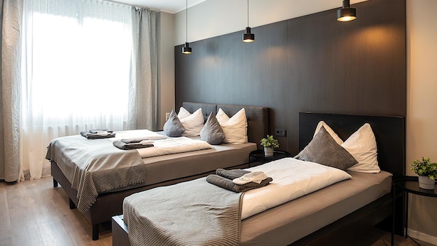Ein modernes Hotelzimmer mit zwei Betten, einem großen Fenster mit Vorhängen und Pendelleuchten.