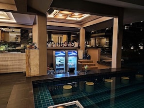 Uma área de bar com uma piscina ao primeiro plano, um espaço de estar com balcão de bar e uma escadaria que leva a um andar superior.