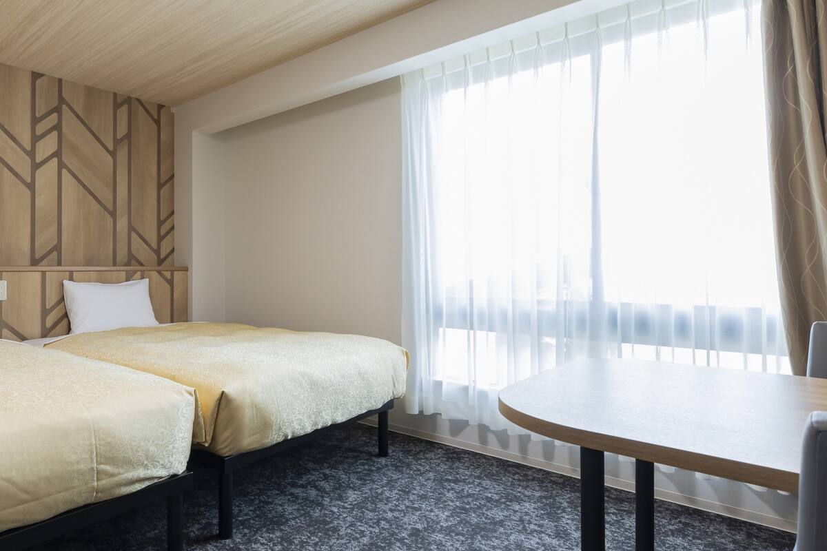 Ein Hotelzimmer mit zwei Betten, einem runden Tisch und einem großen Fenster mit Vorhängen.