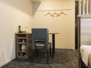 Une chambre d’hôtel de taille réduite, comprenant un lit simple, un petit bureau avec une chaise, une étagère avec des articles de toilette et un porte-manteau avec des cintres.