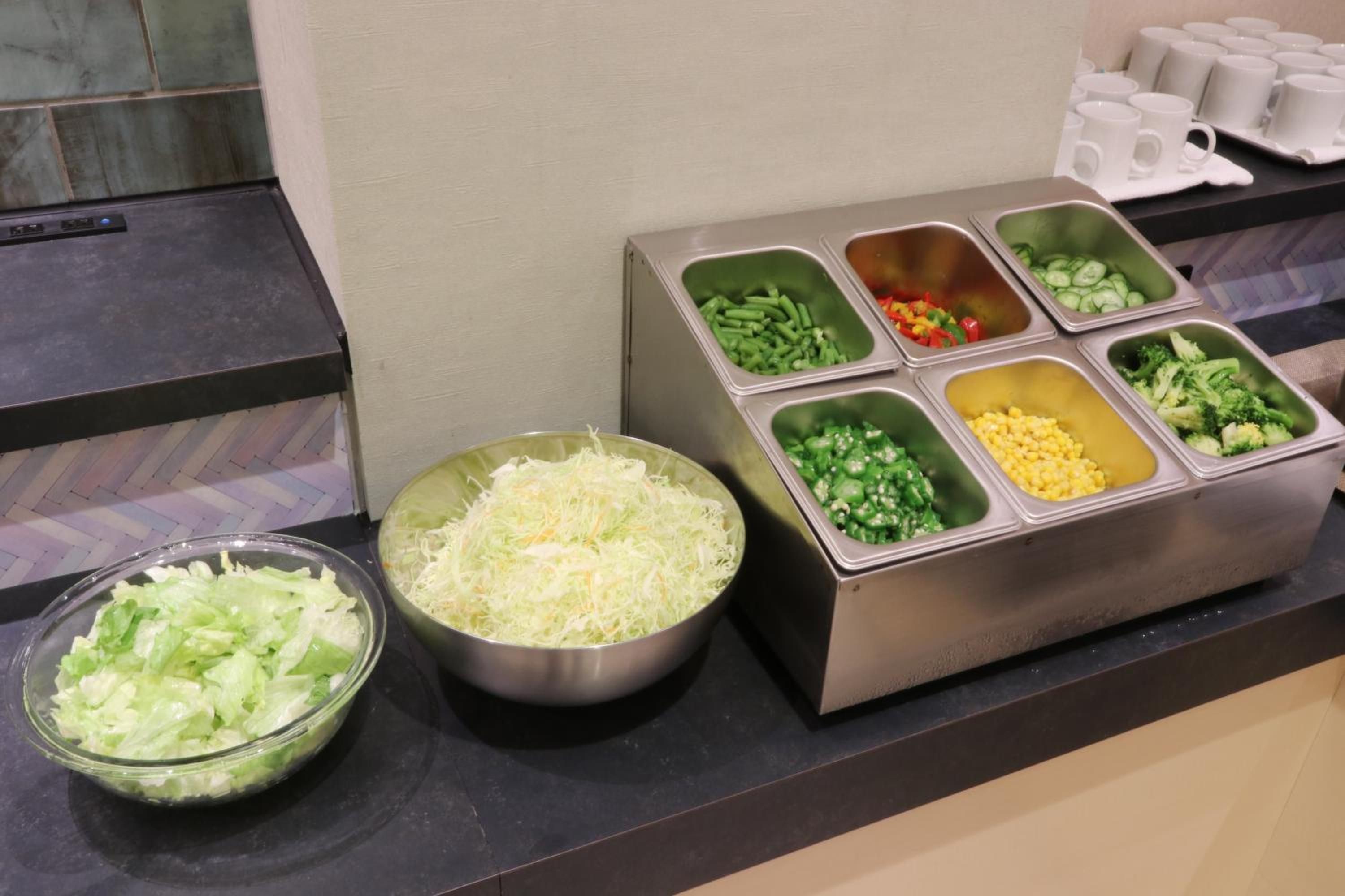 En buffet med forskellige madmuligheder, herunder revne grøntsager, grønne bønner, peberfrugter, broccoli, majs og en skål med salat.