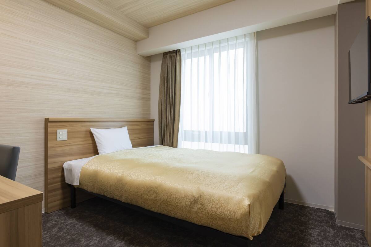 Ein Hotelzimmer mit Holz-Kopfteil, einem Einzelbett mit beiger Bettdecke, einem Schreibtisch mit Stuhl, einem Fenster mit transparenten Vorhängen und einem an der Wand montierten Flachbildfernseher.