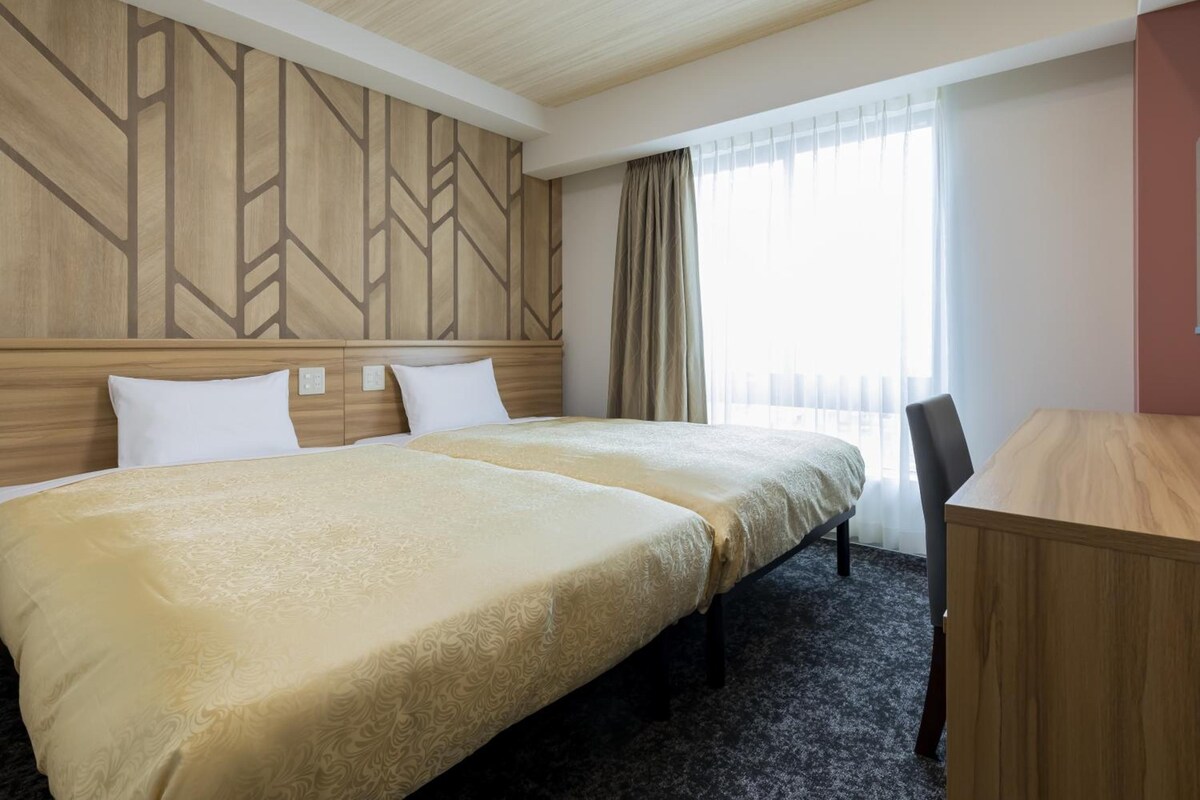 Ein Hotelzimmer mit zwei Betten, einem Schreibtisch und einem fensterbepflanzten Fenster.