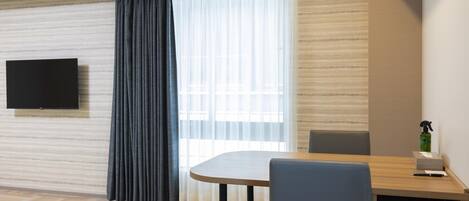Habitación moderna con televisión de pantalla plana en la pared, una mesa de comedor con sillas, un ventanal con cortinas translúcidas y un escritorio pequeño con una computadora portátil y un atomizador.