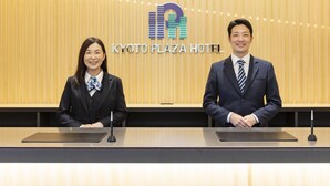 Two individuals seated at a reception desk with a wooden backdrop and a hotel logo. - Kyoto Plaza Hotel Kintetsu Jujo (Kyoto)