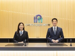 Two individuals seated at a reception desk with a wooden backdrop and a hotel logo. - Kyoto Plaza Hotel Kintetsu Jujo (Kyoto)