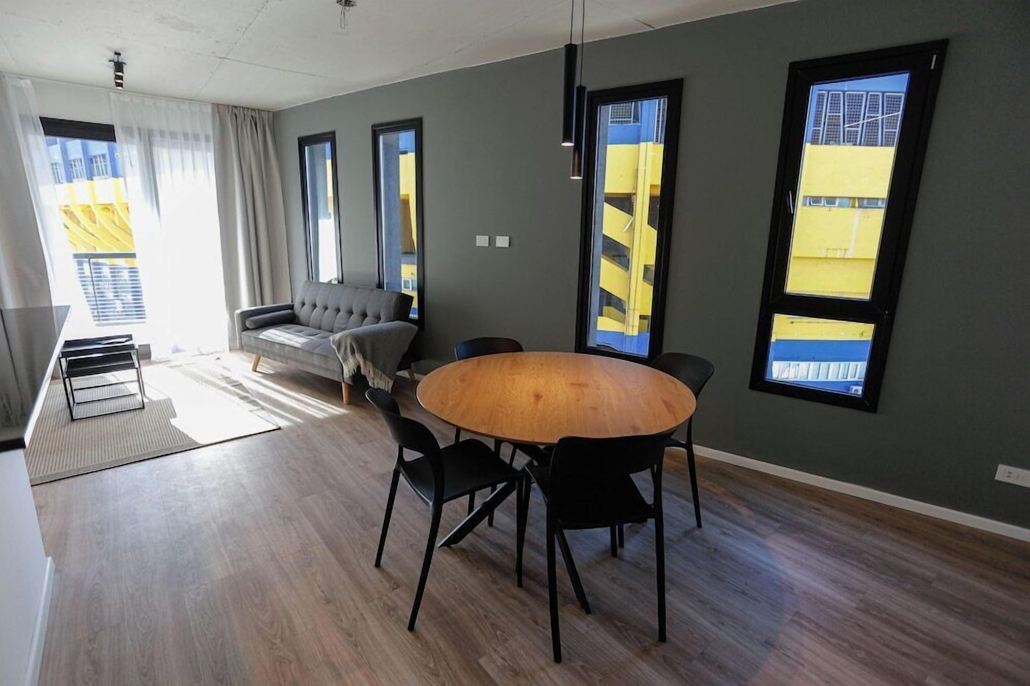 Una sala de estar moderna con una mesa redonda de madera, sillas negras, un sofá gris y grandes ventanas con vistas a edificios amarillos.