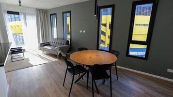 Una sala de estar moderna con una mesa redonda de madera, sillas negras, un sofá gris y grandes ventanas con vistas a edificios amarillos.