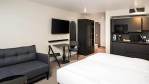 A modern hotel room with a sofa, a dining table, a TV, a wardrobe, and a kitchenette. - ANA Living Karlsruhe, Trademark Collection by Wyndham (Karlsruhe)