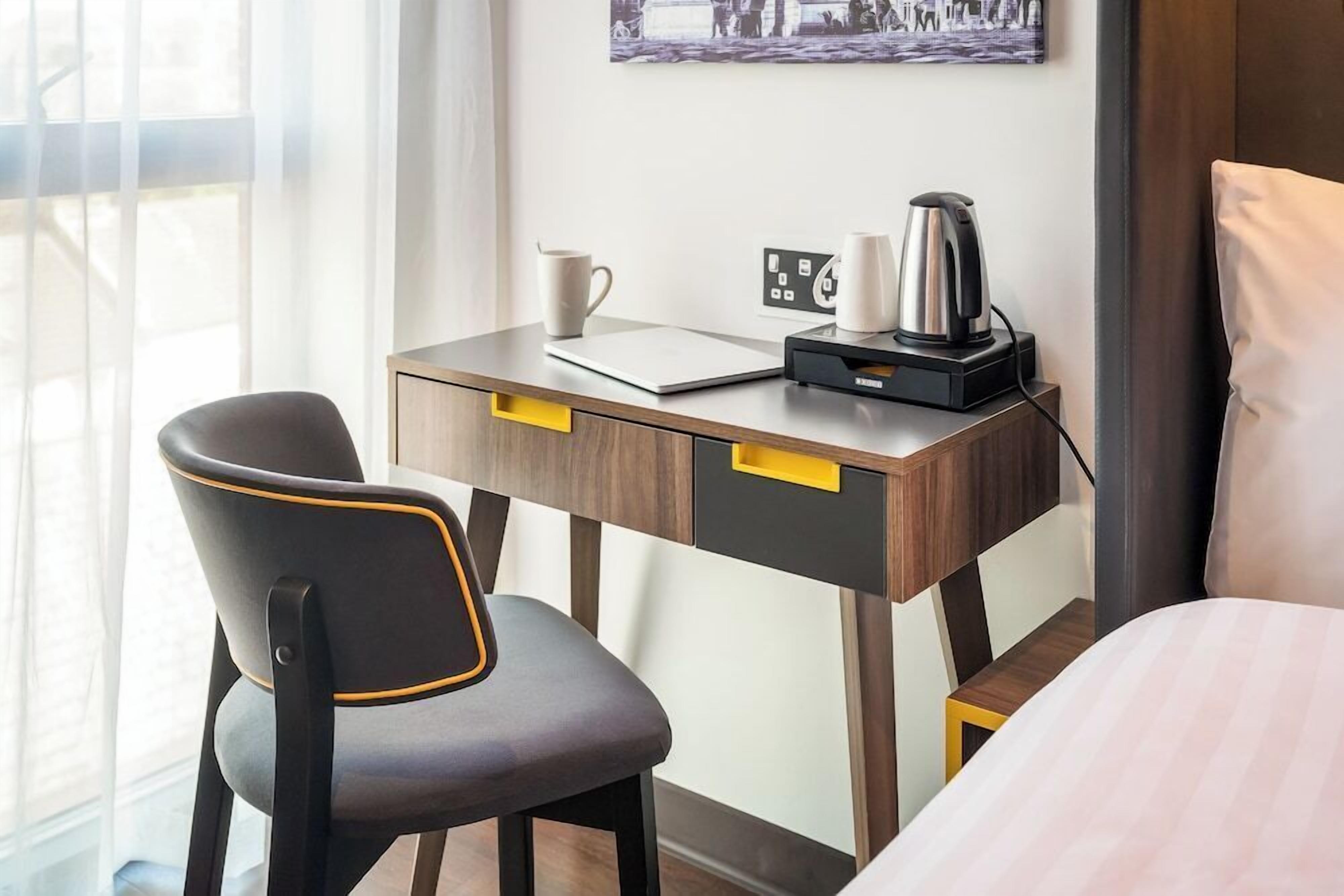 A hotel room with a desk, a chair, a bed, a coffee maker, and a wall-mounted power outlet.