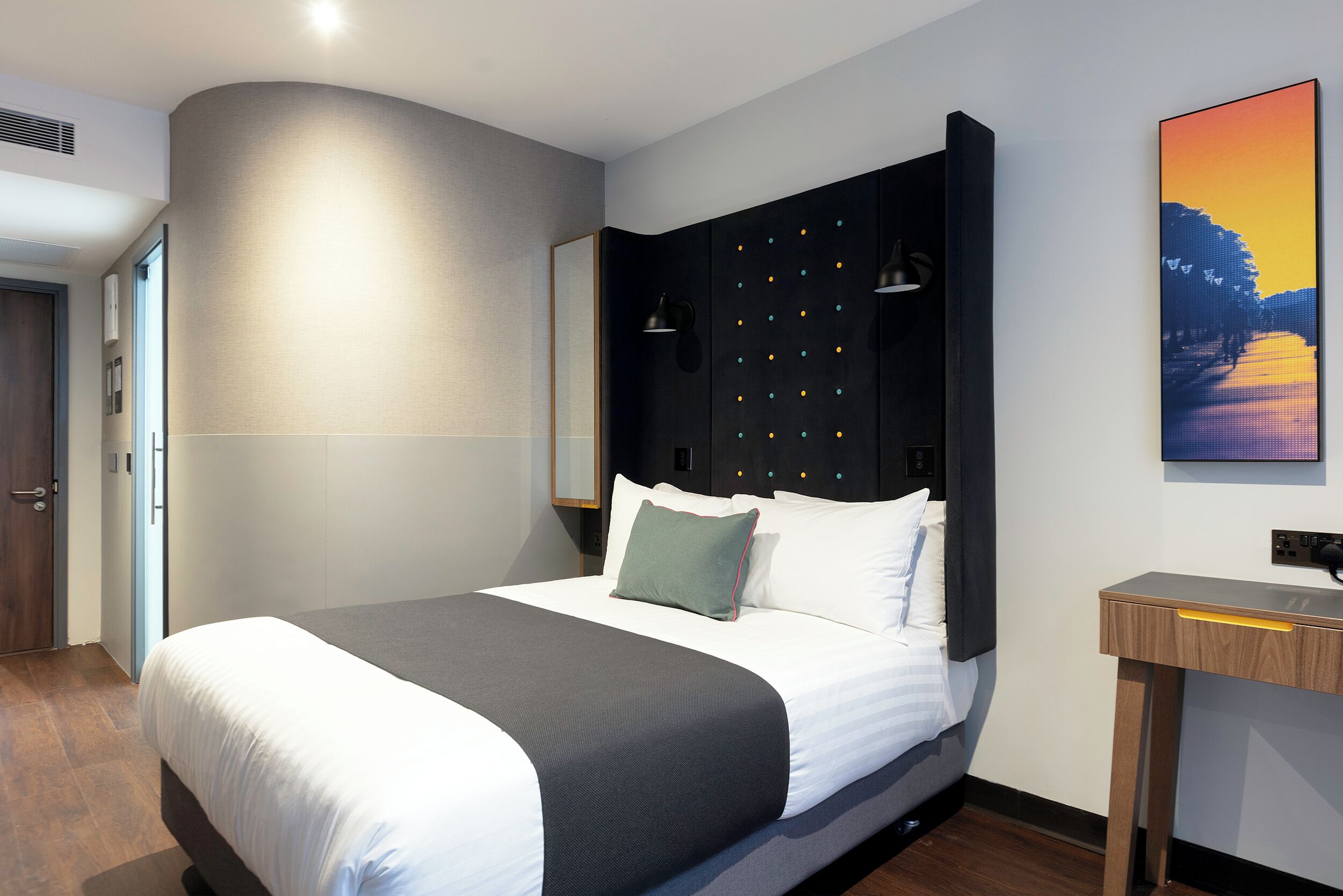 A modern hotel room with a large bed, a dark headboard, a bedside table, and a wall-mounted TV.