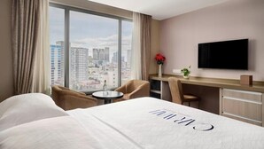 A hotel room with a bed, a desk, a chair, a TV, and a large window with a city view. - Kravan (Phnom Penh)