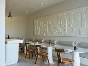 A modern dining area with round tables, wooden chairs, and a decorative wall piece. - Ark Santorini (Santorini)