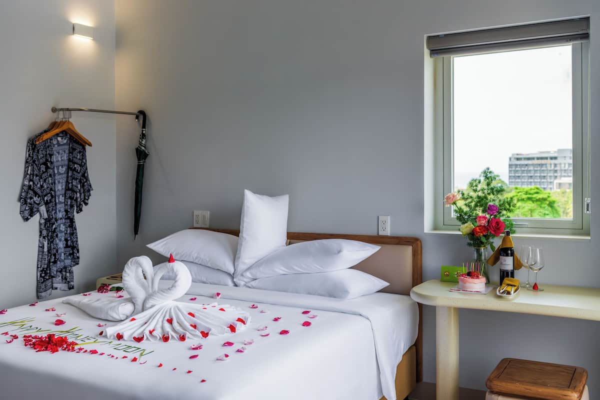 A hotel room with a neatly made bed, a window with a view, a bedside table with a vase of flowers, and a small wooden stool.