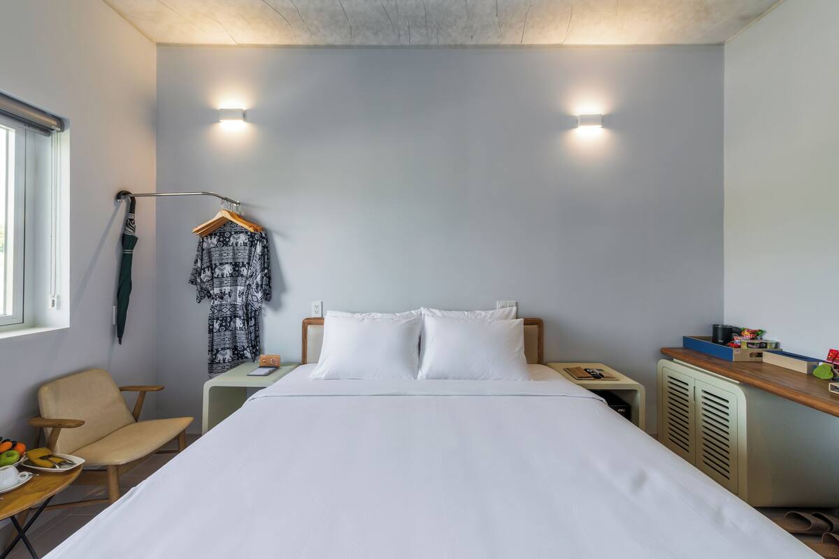 A hotel room with a bed, a chair, a desk, a coat rack, and a nightstand.