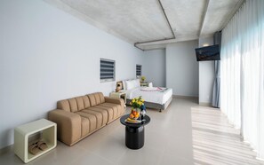 A modern hotel room with a sofa, a small side table, a coffee table with fruit and glasses, a bed with a white bedspread, and a TV mounted on the wall. - The Up Phu Quoc (Phu Quoc)