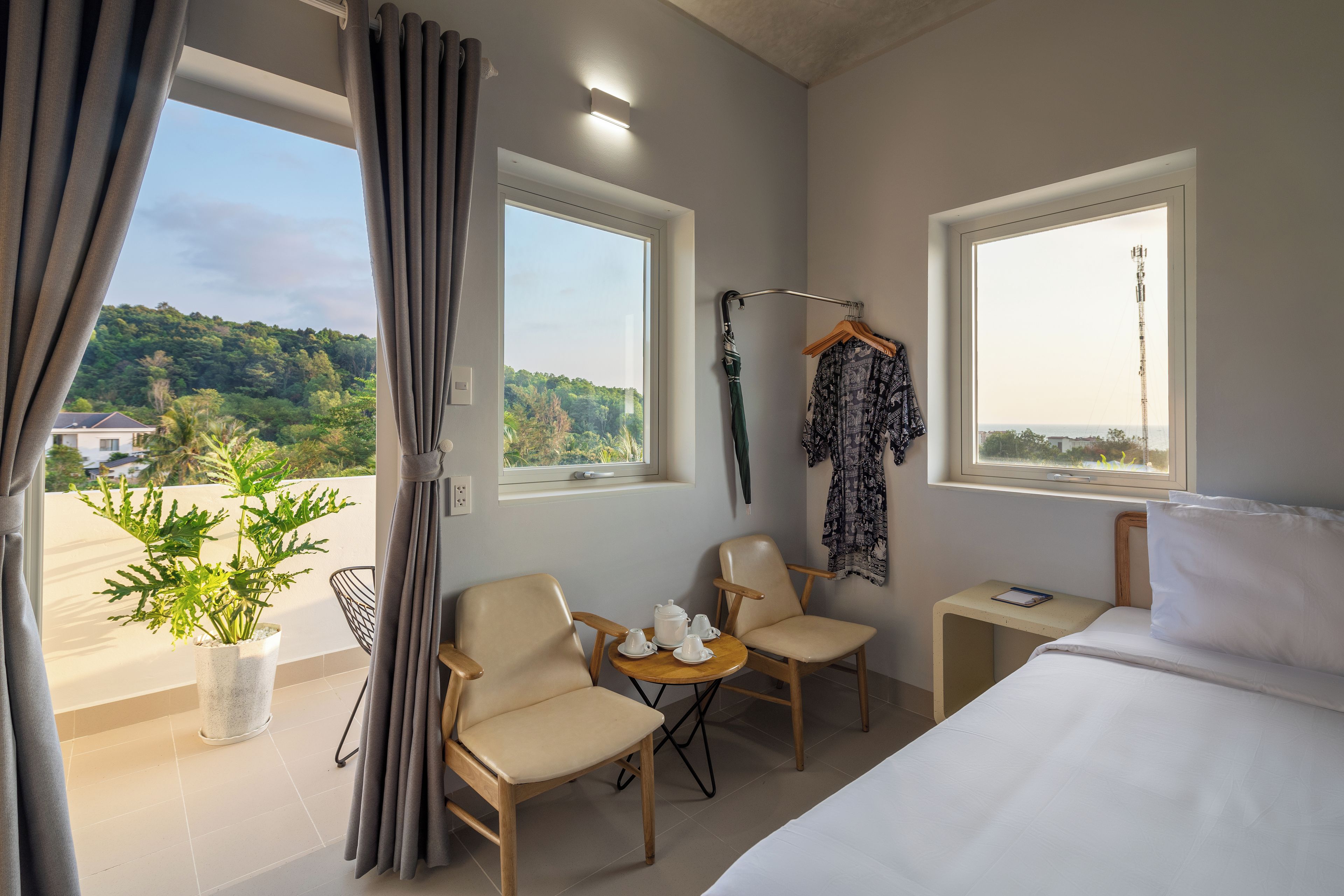 A hotel room with a bed, two chairs, a small table with a teapot, a plant, and a view of greenery outside.