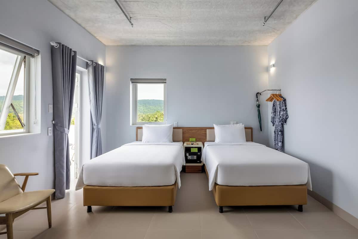 A hotel room with two beds, a chair, a nightstand, and a view of greenery.