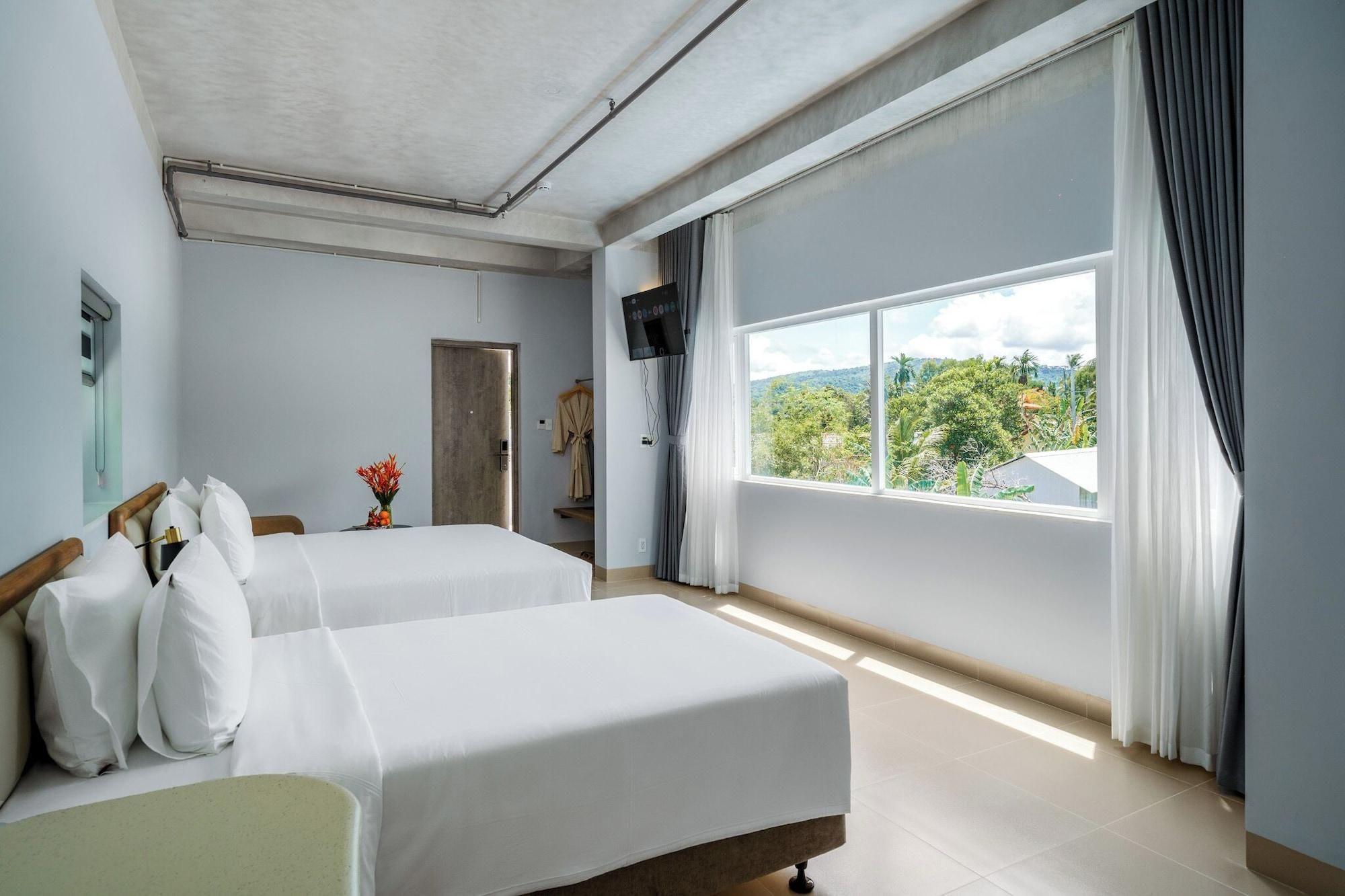 A hotel room with two beds, a TV, a window with a view of greenery, and a door.