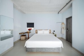 A modern hotel room with a large bed, a bedside table, a mirror, a window with blinds, a robe hanging on a hook, and a door. - The Up Phu Quoc (Phu Quoc)
