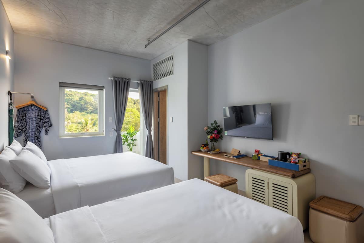 A hotel room with two beds, a TV, a desk with a plant, and a window with curtains.