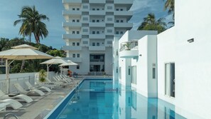 A swimming pool with sun loungers and umbrellas, a white building with balconies, and palm trees in the background. - The Up Phu Quoc (Phu Quoc)