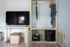 Une chambre équipée d’une télévision, d’un petit meuble avec un coffre-fort, d’un banc et d’un porte-manteau avec une veste.