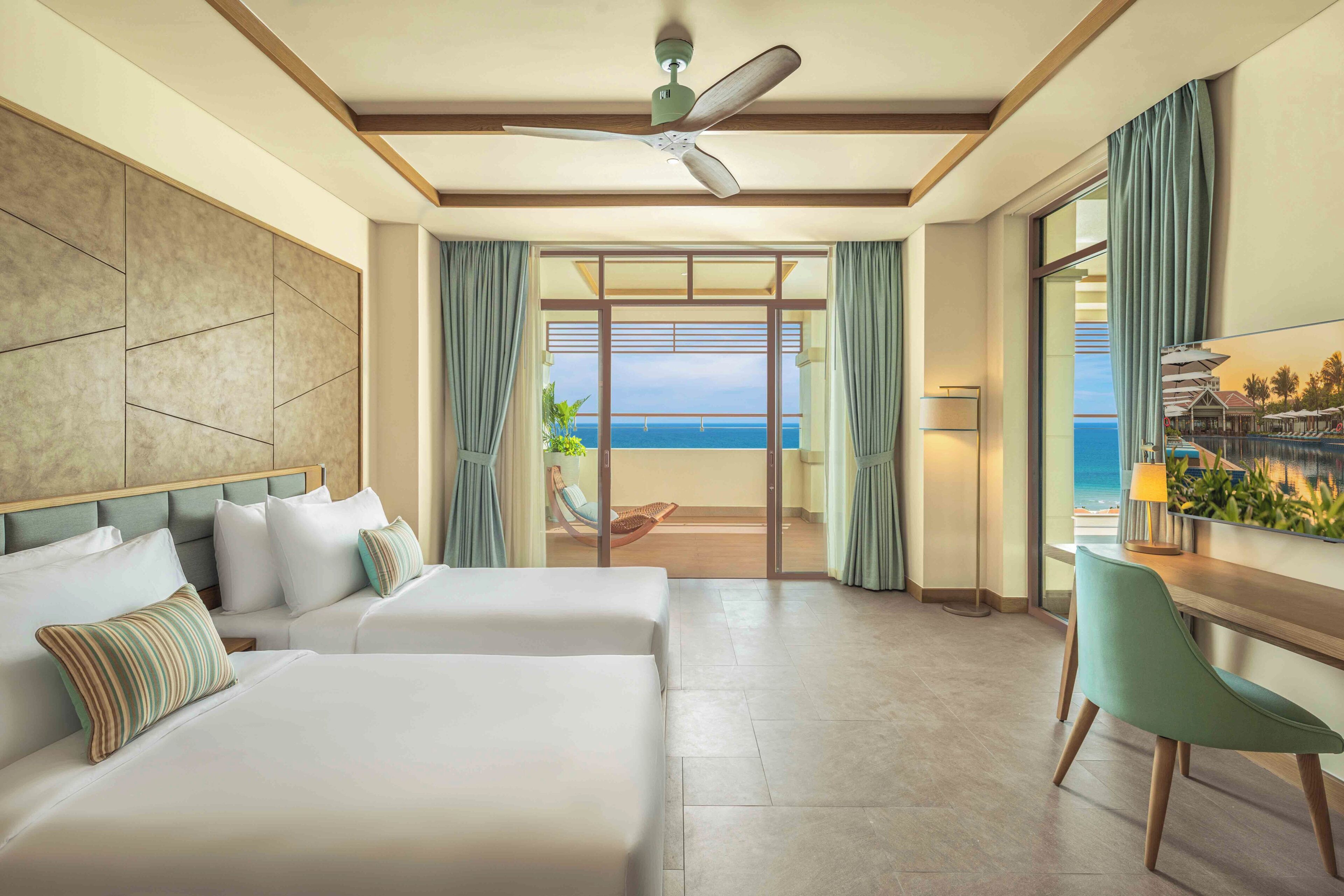 A hotel room with two beds, a desk, a chair, a ceiling fan, and a view of the ocean.